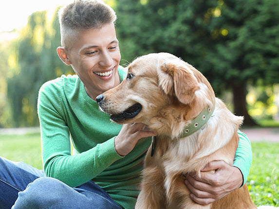 A man sits outdoors on grass, petting a golden retriever.